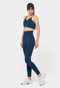 Jūras zilas sporta krūšturis ar platu priekšpusi un saskaņotas augsto vidukļa leggingi, kas aprīkoti ar kabatu un logotipu, izgatavoti no teksturēta materiāla.