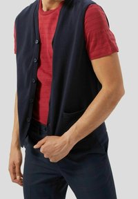 Námořnický pletený svetr s předními knoflíky a kapsou, vrstvený přes červené pruhované tričko. Nosené s tmavě kostkovanými kalhotami.