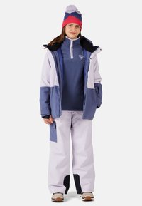 Lyžařský outfit zahrnující námořnickou modrou a bílou bundu, světle fialové kalhoty, pletenou čepici v růžové a modré barvě s bambulí a hnědé akcentové boty.