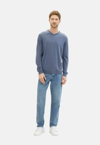 Maglione azzurro a V, maniche lunghe, texture morbida, abbinato a jeans azzurri chiari e sneakers bianche. Il modello è in piedi su uno sfondo semplice.