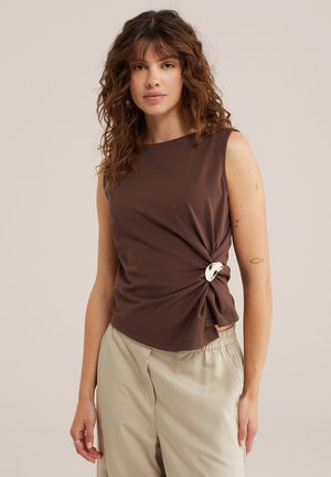 Femme aux cheveux bruns bouclés portant un haut marron sans manches avec un détail de boucle argentée et un pantalon beige à fines rayures, debout devant un fond neutre.