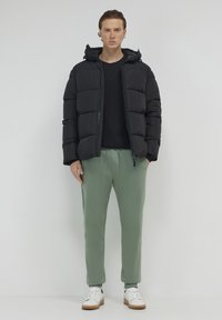 Schwarze Steppjacke mit Kapuze, gestepptem Design, kombiniert mit hellgrünen Jogginghosen und weißen Sneakers mit braunen Akzenten.