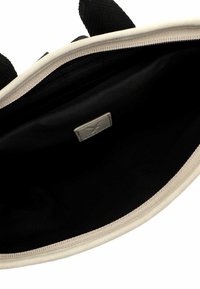 Ylhäältä katsottuna avoin beige-musta laukku, jossa näkyy tilava musta sisusta ja pieni suorakaiteen muotoinen etiketti sisällä.