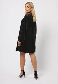 Schwarzes Velourskleid mit langen Ärmeln, lässiger Form und rundem Ausschnitt. Getragen mit schwarzen Absatzsandalen, die die glatte Textur zur Geltung bringen.