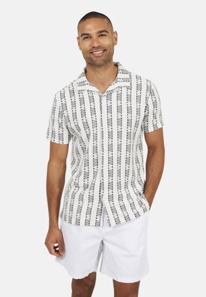 Uśmiechnięty mężczyzna z krótkimi włosami, ubrany w białą koszulę na krótki rękaw z czarno-białym wzorem oraz białe szorty, stojący z jedną ręką w kieszeni.