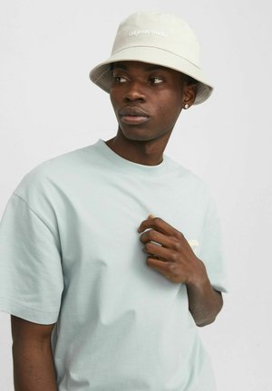 Hellblaues Baumwoll-T-Shirt mit kurzen Ärmeln, auf dem ein dezentes Logo zu sehen ist. Kombiniert mit einem beigen Bucket Hat, der ein schlichtes Design und Schriftzug hat.
