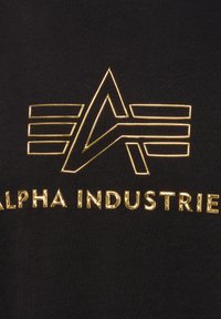 Črn material z zlatim logotipom, ki vključuje stilizirane črke "A" in "I" s horizontalnimi črtami ter teksturiranimi poudarki spodaj.