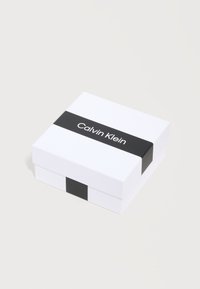 Λευκό τετράγωνο κουτί με μαύρη ταινία που φέρει το κείμενο "Calvin Klein" στην κορυφή, τοποθετημένο σε απλό φωτεινό φόντο.