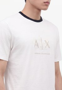 Άνδρας που φοράει λευκό T-shirt Armani Exchange με μαύρο γιακά και κεντημένο λογότυπο στο στήθος, εμφανιζόμενος από τον αυχένα έως τη μέση.