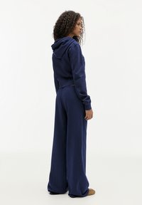 Donna con capelli ricci che indossa una felpa blu navy e pantaloni larghi, in piedi di lato su uno sfondo bianco.