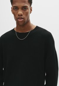 Junger Mann mit kurzen lockigen Haaren, der einen schwarzen strukturierten Pullover, eine silberne Halskette und kleine Ohrringe trägt, vor einem schlichten hellen Hintergrund.