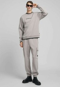 Felpa grigia con scritte nere, abbinata a pantaloni cargo coordinati. Entrambi hanno una vestibilità comoda e tessuto strutturato. Indossati con stivali neri.