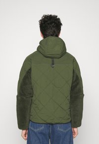 Veste matelassée verte avec une capuche, présentant des manches texturées et duveteuses d'un vert foncé et un détail de resserrement à l'arrière. Coupe ajustée, avec des coutures visibles.