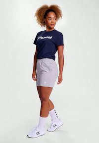 T-shirt azul-marinho com logótipo branco, calções cinza com cintura elástica, meias brancas até ao joelho e sapatilhas pretas. Tecido liso e ajuste descontraído.