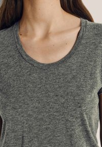 Moteris dėvi pilką marškinėlių su apvalia iškirpte, arti matomi kaklo, raktikaulio ir viršutinės krūtinės dalys, matyti ilgų rudų plaukų sruogos.