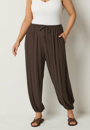 Vrouw draagt donkerbruine losse broek met enkelboorden, wit mouwloos topje en bruine sandalen, staand met één hand in haar zak.