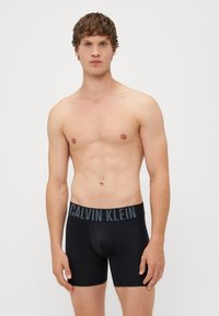 Boxeri negri dintr-un material neted, cu un elastic lat pe talie inscripționat cu logo-ul "Calvin Klein" în gri. Design ajustat, cu lungime până la mijlocul coapsei.