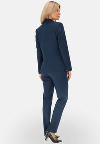 Granatowy garnitur skrojony na miarę, z dopasowaną marynarką i prostymi spodniami, wykonany z gładkiego materiału. Czarne szpilki z ozdobnym akcentem w postaci kokardy.