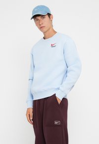 Light blue sweatshirt with red Nike logo and text; paired with deep burgundy pants featuring a white Nike patch. Casual athletic design.