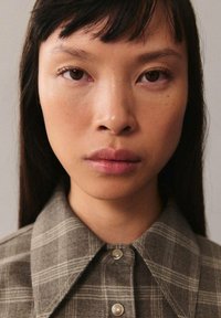 Kauluspaita, jossa on harmaasävyinen ruudullinen kuvio, tekstuurilla varustettu kangas ja nappikiinnitys kaulassa. Sileä iho, tumma hius.