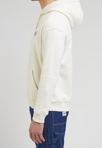 Sweat à capuche crème avec poche kangourou, capuche à cordon, poignets et ourlet côtelés ; texture douce et détail de marque minimal à l'avant.