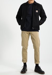 Giacca nera con zip e tasca sul petto, felpa a motivi blu navy sotto, chinos beige arrotolati e scarpe nere.