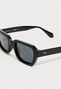 Mustat suorakulmaiset aurinkolasit paksuilla kehyksillä, tummilla linsseillä ja hienovaraisella kultaisella yksityiskohdalla tempurin varressa, esitettynä vaalealla taustalla.