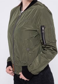 Bomberka w oliwkowym zielonym kolorze z błyszczącym nylonowym wykończeniem, ściągacze przy mankietach i kołnierzu, zamek błyskawiczny z przodu oraz kieszeń na zamek błyskawiczny na ramieniu.