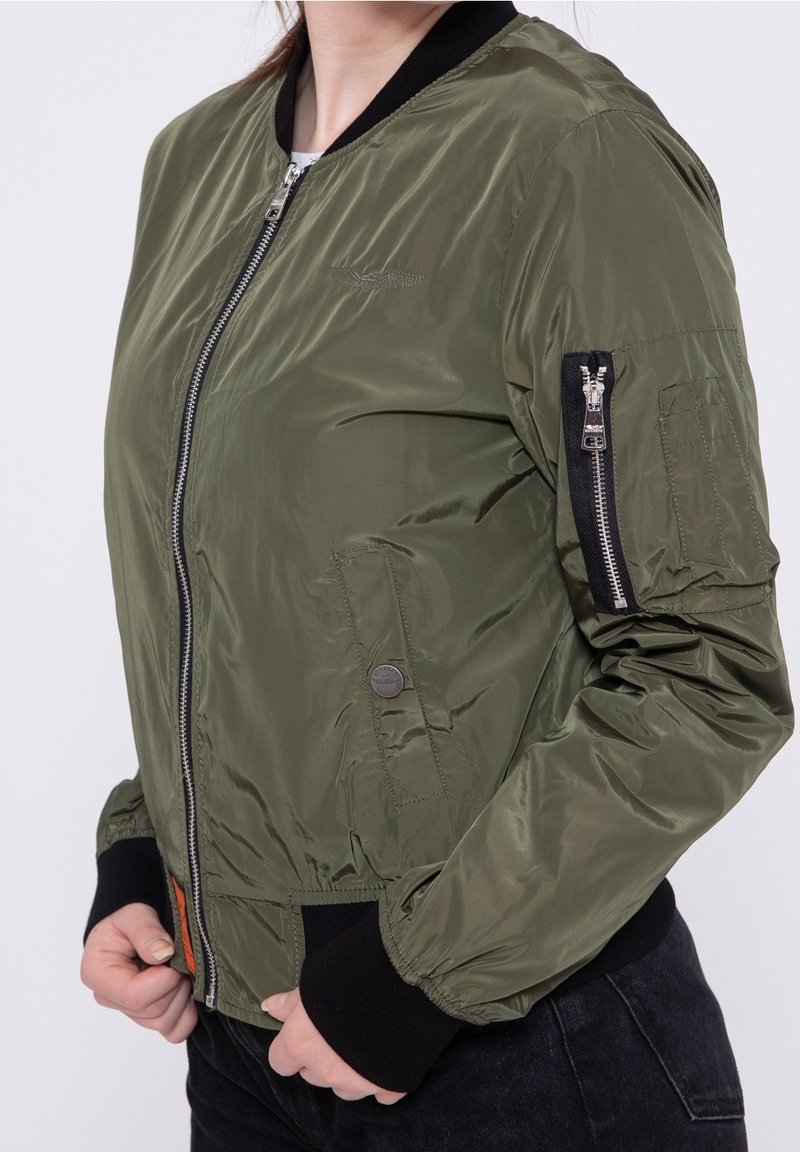 Bomberka w oliwkowym zielonym kolorze z błyszczącym nylonowym wykończeniem, ściągacze przy mankietach i kołnierzu, zamek błyskawiczny z przodu oraz kieszeń na zamek błyskawiczny na ramieniu.