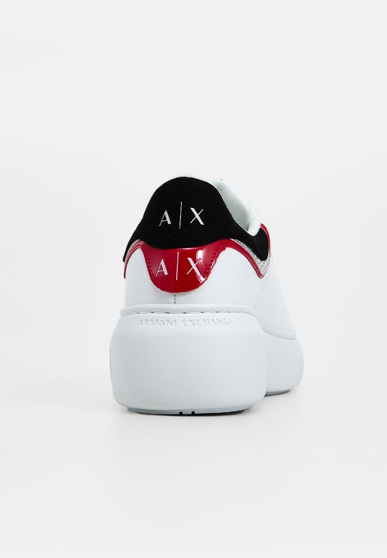 Białe sneakersy o zaokrąglonym kształcie, z czarnymi i czerwonymi akcentami, zamszowym paskiem na pięcie oraz wytłoczonym logo marki na pięcie i podeszwie.
