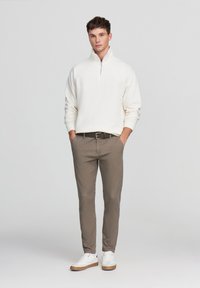 Maglione bianco con cerniera abbinato a pantaloni chinos marrone chiaro e sneaker bianche. Il maglione ha un colletto alto e polsini a coste.