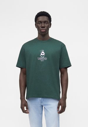 Fiatal férfi sötétzöld pólóban, amelyen egy kávéscsésze grafikája és a „Coffee Party Life begins after coffee” felirat látható, világoskék farmerrel párosítva.