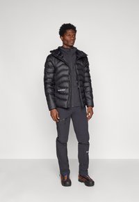 Black puffer jacket with a hood, zip front, and logo; paired with dark gray pants and textured, multi-colored hiking shoes.