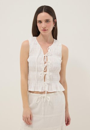 Mujer joven con cabello castaño liso que lleva una blusa blanca sin mangas con fruncidos atada al frente y una falda blanca, sobre un fondo liso.