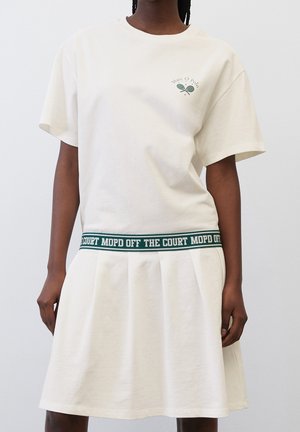Personne portant une robe blanche à manches courtes avec une ceinture verte portant l'inscription "OFF THE COURT MOPD" et un petit logo vert de raquette de tennis sur la poitrine.