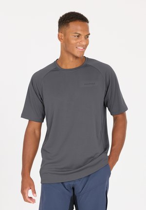 Urheilullinen mies, joka pukeutunut tummanharmaaseen lyhythihaiseen urheilupaitaan ja sinisiin shortseihin, seisoo käsi taskussa ja katsoo sivulle hymyillen.