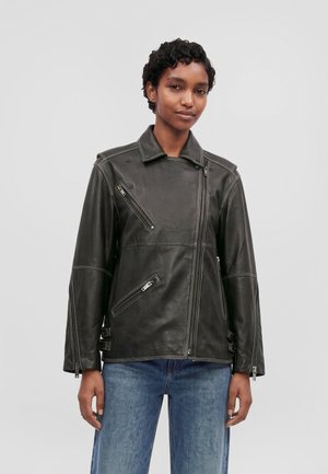 Un mannequin aux cheveux courts et bouclés porte un blouson de motard en cuir noir à fermetures éclair et un jean bleu, sur un fond uni clair.