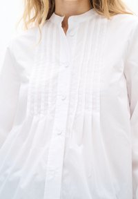 Blouse blanche à manches longues avec plis verticaux sur le devant, patte boutonnée et encolure sans col portée par une personne aux cheveux blonds.