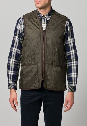 Gilet matelassé vert olive avec fermeture éclair sur le devant, poches latérales et bord marron. Porté par-dessus une chemise à carreaux bleu et blanc, mettant en valeur un style superposé.