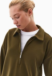 Felpa zip-up verde oliva con colletto, caratterizzata da una cerniera argentata, texture liscia e indossata sopra una camicia bianca.