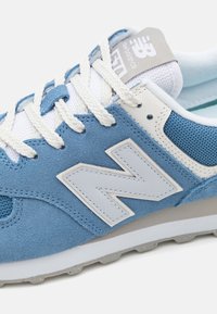 Ljust blå mocka- och mesh-sneakers med vita accenter, prydda med en framträdande vit "N"-logotyp och strukturerade snören på en grå gummisula.