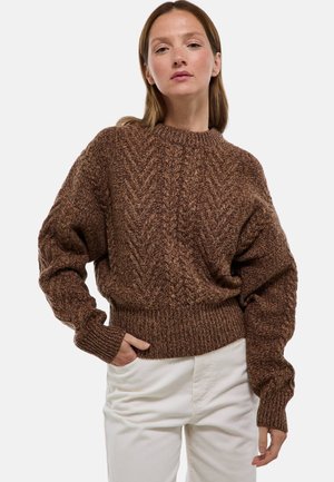 Pull en maille marron avec un motif en chevrons, des poignets et un ourlet côtelés, et une coupe décontractée. Porté avec un pantalon en velours côtel�é blanc.