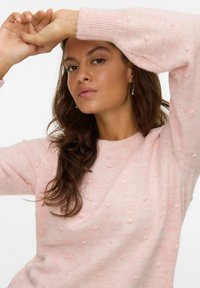 Ženska z dolgimi temnimi lasmi, oblečena v svetlo rožnato strukturno pulover, dviga roke in gleda neposredno naprej na enobarvni ozadju.