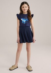 Mörkblå t-shirt med volanger på ärmarna och en valgrafik, tillsammans med en texturerad mörkblå kjol och beige slip-on skor.