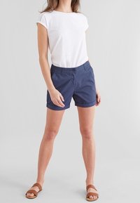 White short-sleeve top with subtle texture, paired with navy blue rolled-up shorts and brown flat sandals. Simple and casual design.