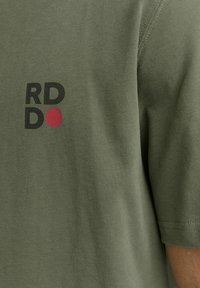 Camiseta verde oliva de manga corta, con un gráfico en negro y rojo que dice "RD DO" impreso en la parte superior izquierda. Textura de tela suave.
