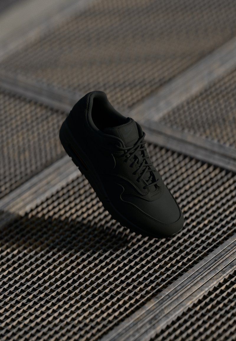Sneaker preto com uma textura suave, biqueira redonda, atacadores à frente e uma marcação sutil. Exibido contra um fundo industrial texturizado.
