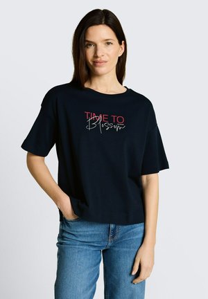 Donna con capelli castani lisci che indossa una maglietta nera con la scritta "TIME TO Blossom" e jeans blu, in piedi con una mano in tasca.