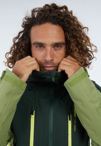 Nepremokavá tmavozelená bunda so svetlozelenými akcentmi, vysokým golierom a nastaviteľnými manžetami. Obsahuje vertikálne zipsy a textúrovaný povrch.