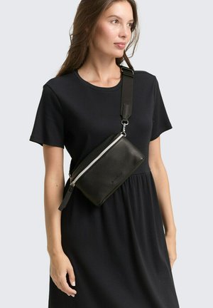 Borsa a tracolla in pelle nera con superficie texture, zip in argento e tracolla regolabile. Indossata su un vestito nero.